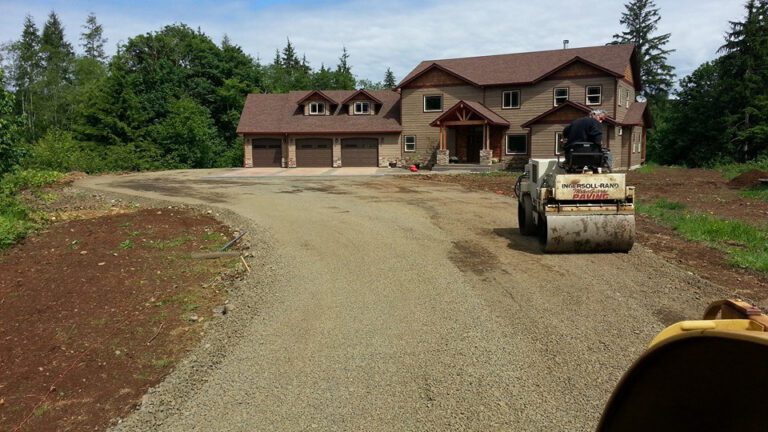 Worker driving road roller on dirt driveway leading to brown home